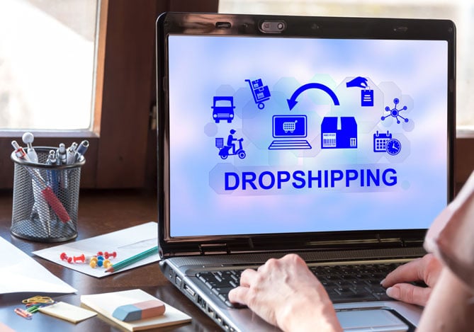 Mercado Pago: pessoa em frente a um notebook, que está com a palavra “dropshipping” e símbolos relacionados ao tema na tela, além de materiais de escritórios espalhados na mesa para aprender como calcular lucro das vendas.