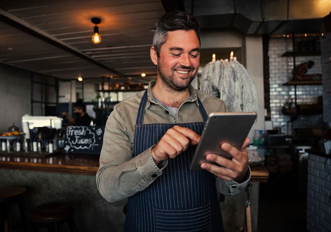 Homem vestindo uma camisa verde com um avental azul listrado, segurando um tablet e em pé em um restaurante. Ele está em frente ao balcão de bebidas e está pesquisando como gerar link de pagamento com cartão de crédito no Mercado Pago. 