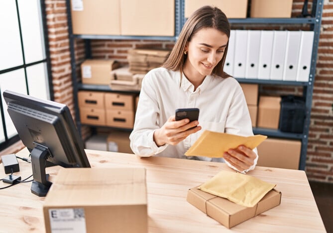 Mercado Pago: Empresária organizando as compras online da sua empresa para a Black Friday. Ela está segurando um celular e uma encomenda, apoiada em uma mesa de trabalho com outras encomendas e um computador.