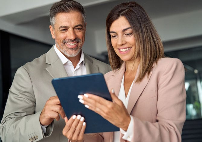 Mercado Pago: imagem de dois sócios de uma empresa realizando uma consulta ao CNPJ por meio de um tablet. Na imagem, é possível ver que a mulher segura o tablet em suas mãos, enquanto o homem toca a tela para que, juntos, identifiquem se há alguma necessidade de atualização de cadastro. Ambos estão sorrindo na foto.