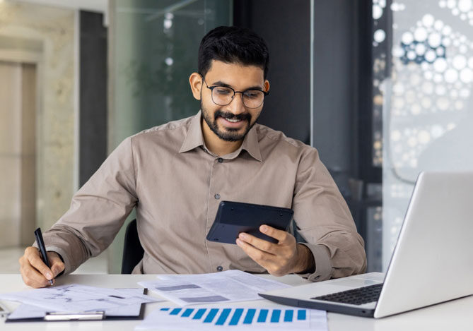 Mercado Pago: imagem de um empresário sentado à mesa de trabalho enquanto realiza o controle financeiro de sua empresa. Sobre a mesa, é possível ver alguns relatórios impressos e um notebook aberto. em uma das mãos, o empresário tem um celular e, na outra, uma caneta, que usa para fazer anotações sobre sua gestão empresarial.