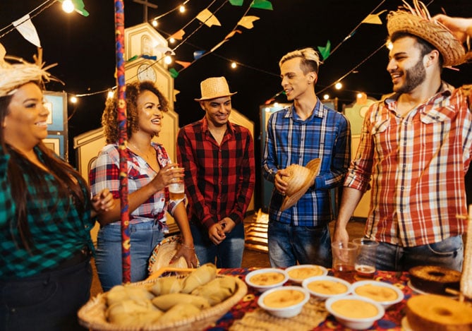 Mercado Pago: grupo de pessoas vestidas de caipiras com camisas xadrez, calça jeans e alguns com chapéu de palha em uma praça com bandeirinhas e em volta de uma mesa com comidas típicas de Festa Junina para empresas
