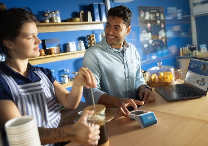 Imagem de empresários em sua cafeteria.  É possível ver a empresária preparando um café com uma prensa francesa, enquanto o empresário utiliza a maquininha Point do Mercado Pago para registrar dados de uma venda a fim de obter informações para serem analisadas posteriormente via aplicativo como uma forma de medir a eficiência operacional da empresa.