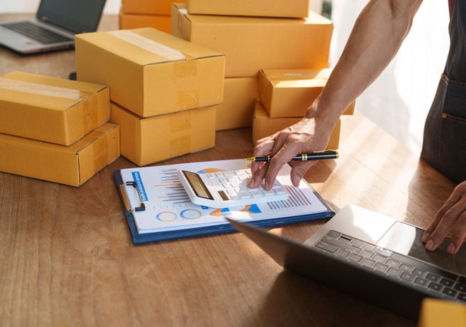 Mercado Pago: mãos mexendo em uma calculadora e um notebook em cima de uma mesa, onde também há caixas e planilhas para fazer a gestão financeira do e-commerce.