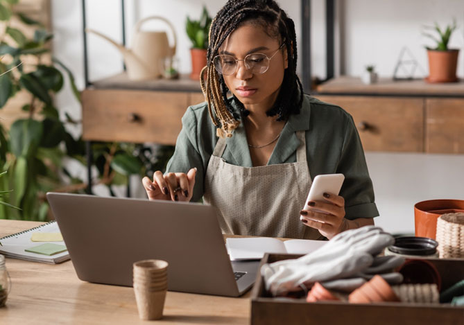 Imagem de uma empresária em seu estúdio de cerâmica sentada à mesa de trabalho. Na imagem, é possível ver a empresária usando um notebook apoiado sobre a mesa, enquanto segura um celular em uma das mãos. Ela está usando o computador para verificar como fazer um estorno de pagamento com a sua maquininha Point Mercado Pago. Sobre a mesa, é possível ver peças de cerâmica, vasos de enfeite e um caderno para anotações.