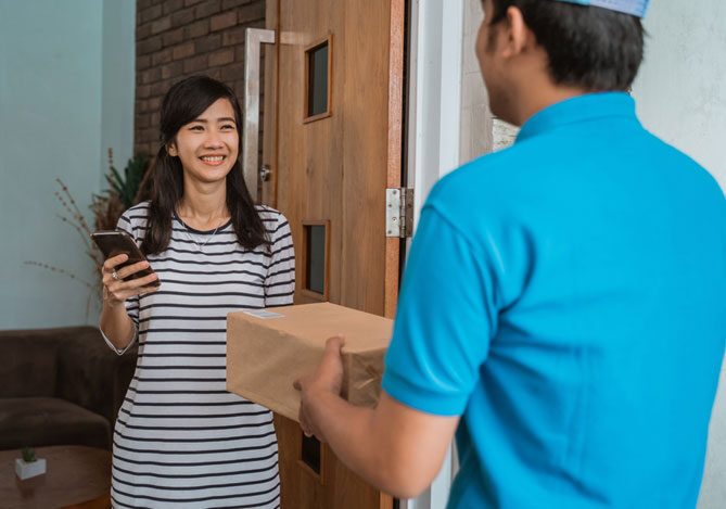 Mercado Pago: imagem de uma mulher sorrindo recebendo o produto que comprou pela internet à porta de sua casa. Na imagem, também é possível ver o entregador vestindo camisa e boné azul, representando um dos fornecedores que faz entregas para lojas de dropshipping. Ele está segurando o pacote para entregar à cliente.