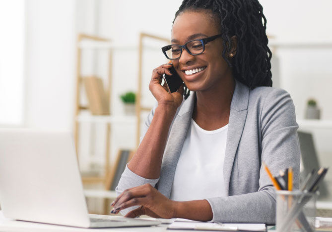 Mercado Pago: imagem de uma empresária sentada à mesa de trabalho enquanto digita em seu notebook e fala ao telefone, demonstrando que está explorando estratégias de vendas ao fazer um follow-up com alguns clientes. Sobre a mesa também é possível ver um porta canetas com algumas canetas e lápis. 