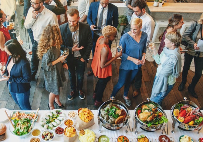 Mercado Pago: imagem de funcionários de uma empresa próximas a uma mesa com diversas opções de comida. As pessoas estão conversando e confraternizando durante um dos eventos corporativos da empresa para celebrar o fim de ano. Na imagem, é possível ver também que algumas pessoas seguram taças com bebidas.