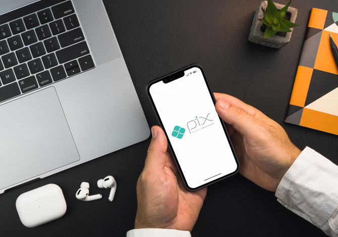 Mercado Pago: imagem das mãos de um empresário segurando um celular com o logo do Pix na tela, indicando que ele está buscando informações de como realizar transações em sua empresa sem que haja a necessidade de pagar a Taxa Pix. O empresário está com as mãos apoiadas sobre sua mesa de trabalho, sobre a qual também é possível ver um notebook, fones de ouvido bluetooth e um pequeno vaso de flor.