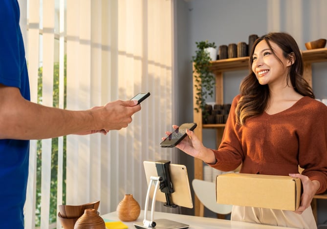 Mercado Pago: mulher segurando uma caixa e mostrando o celular para que o cliente aproxime sua carteira digital conectada ao seu celular para efetuar o pagamento e ter uma experiência do cliente eficiente.