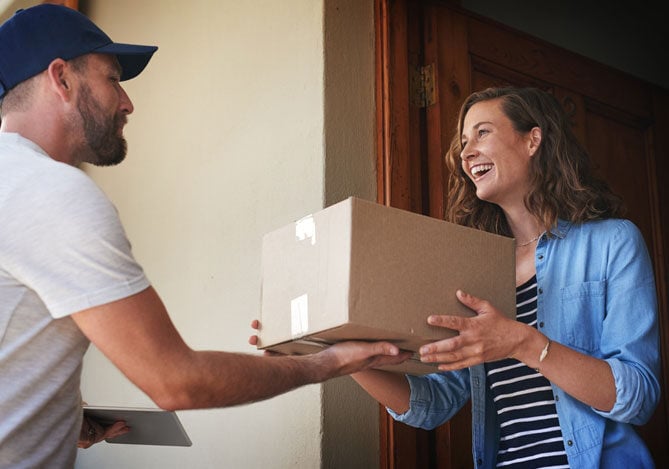 Mercado Pago: imagem de uma consumidora recebendo uma caixa lacrada em sua casa, direto das mãos de um entregador, e sorrindo. A consumidora recebe a caixa por estar em um programa de assinaturas de uma empresa que oferece essa solução como uma estratégia de fidelização de clientes. A imagem ilustra o recebimento dos produtos do mês dessa consumidora.