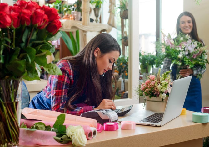 Florista analisando o fluxo de caixa na frente do notebook em sua loja, cercada por flores e materiais de trabalho, enquanto outra funcionária prepara um buquê ao fundo.