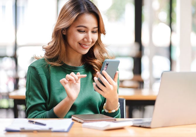 Mercado Pago: imagem de uma empresária sentada à mesa de trabalho usando seu celular para calcular taxas ao cobrar usando link de pagamento. Sobre a mesa também é possível ver um notebook aberto, uma prancheta, uma calculadora e um caderno.