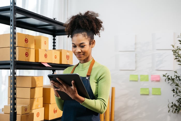 Mercado Pago: mulher em centro de distribuição, ao lado de uma prateleira com caixas e com um tablet na mão para avaliar o processo da logística da sua empresa. 