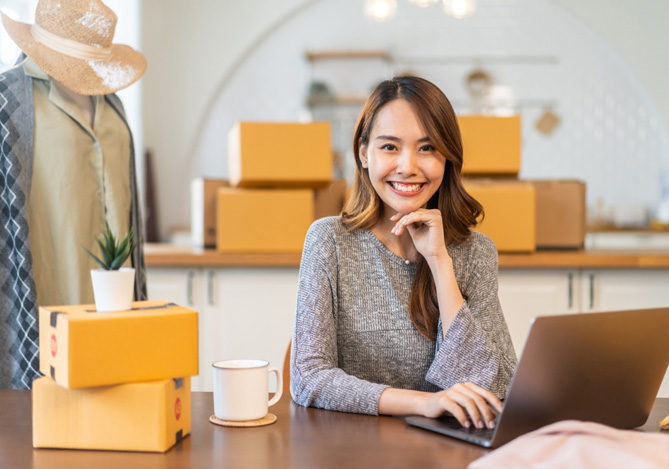 Mercado Pago: mulher sentada à mesa com computador, uma xícara e algumas caixa enquanto monta uma estratégia de promoção de inverno para seu e-commerce