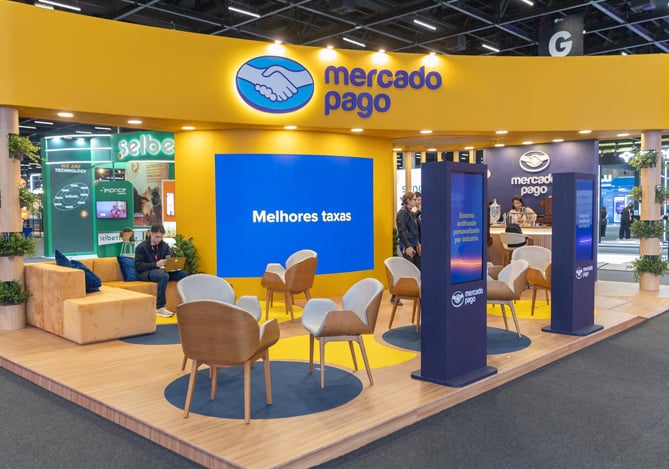 Imagem do estande do Mercado Pago no VTEX Day 2025 com a nova cor do branding, amarelo e azul, com piso de madeira, cadeiras em madeira e outros detalhes com totens e telão exibindo mensagens sobre o ecossistema