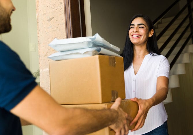 Mercado Pago: imagem de uma mulher sorrindo na porta de sua casa recebendo duas caixas e dois pacotes. Os produtos estão sendo repassados em mãos para a mulher por um entregador que está levando os pedidos que essa cliente realizou em uma empresa após selecionar, entre as diferentes opções de modelos de assinatura, a que melhor atendia suas necessidades para receber pacotes em casa mensalmente, sem precisar ir até uma loja ou realizar pagamentos a cada compra, pois utiliza um pagamento recorrente como forma de pagamento.