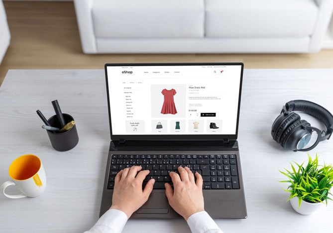 Mercado Pago: Pessoa com mãos no notebook apoiado em mesa, pesquisando sobre o que é review para avaliar uma roupa que comprou através de um dropshipping. Na tela do computador, há a página de compra da roupa e na mesa, há um vaso de planta, um fone preto, um porta-lápis preto e uma caneca branca e laranja. 