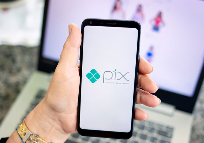 Mercado Pago: Mão de mulher segurando celular com símbolo do Pix, procurando como proteger sua empresa do Pix falso, com notebook aberto ao fundo em página de compra, apoiado em mesa.