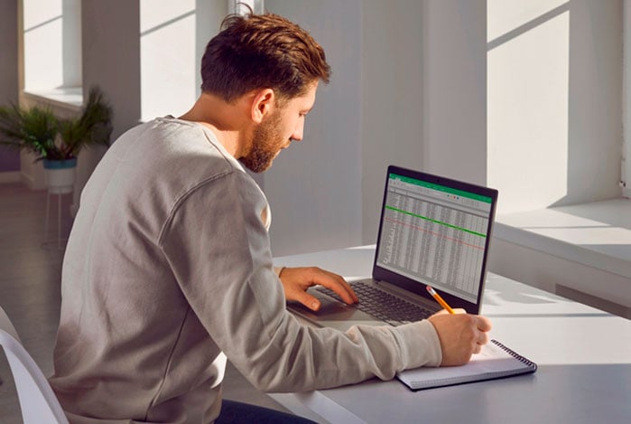 Mercado Pago: homem sentado com computador aberto em cima da mesa, mexendo em uma planilha controle financeiro para empresa, a fim de organizar finanças e em outra mão anotando informações em um caderno.