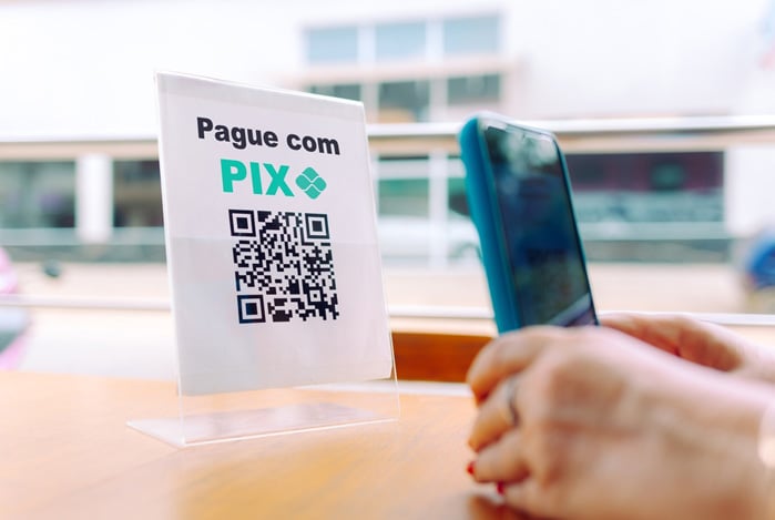 Mercado Pago: mãos segurando um celular apontando para um QR Code Pix CNPJ impresso em uma plaquinha.