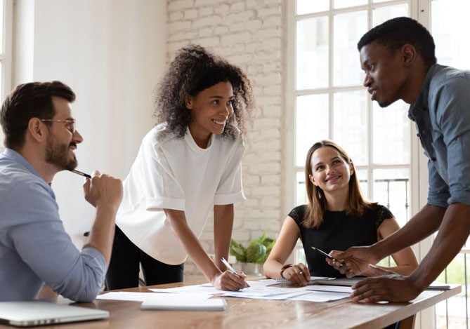 Mercado Pago: imagem de quatro pessoas em volta de uma mesa de reuniões, duas mulheres e dois homens vestindo camisa azul, camiseta branca, camiseta preta e camisa cinza, conversando sobre contratação de força de trabalho enquanto calculam quanto custa um funcionário para a empresa. Sobre a mesa é possível ver alguns documentos com registros e números da empresa.