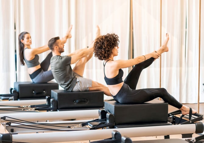 Mercado Pago: três pessoas em um studio de pilates fazendo alongamento de perna em cima de um aparelho com almofada preta.