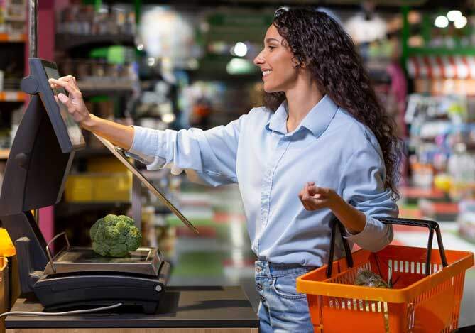 Mercado Pago: Mulher em pé usando camisa azul clara, calça jeans e segurando uma cesta vermelha de supermercado, utilizando um totem de autoatendimento para finalizar um pagamento.