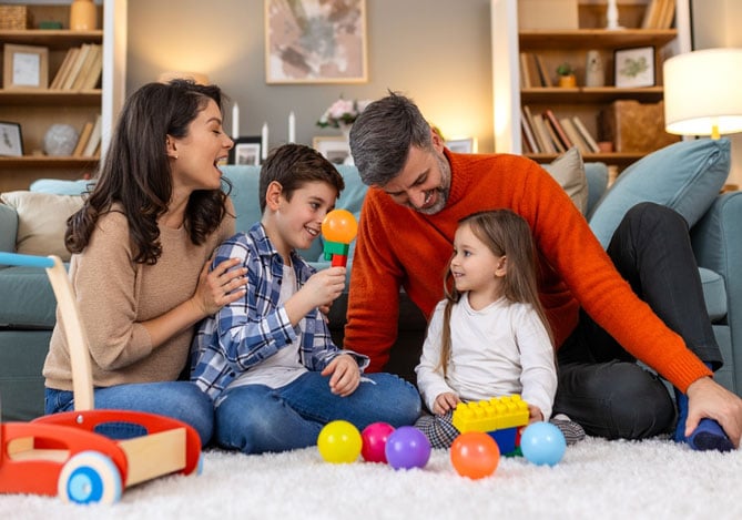 Mercado Pago: Família sentada em tapete na sala de casa com brinquedos de Dia das Crianças. Mulher veste uma blusa bege e calça jeans, menino veste uma camisa xadrez azul e calça jeans, homem veste uma camisa vermelha e calça preta e menina veste uma camisa branca e calça colorida. Atrás está um sofá verde com almofada, na frente há um carrinho de madeira e bolinhas de plástico e todos estão na sala de estar. 