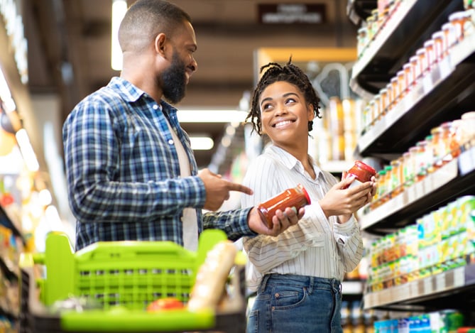 Imagem de um casal sorridente escolhendo alimentos em uma prateleira, ilustrando o comportamento do consumidor, com destaque para o uso do Mercado Pago como método de pagamento.