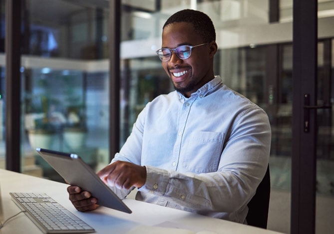Mercado Pago: homem negro sorrindo de óculos vestindo camisa social branca em um escritório com mesa corporativa e computador de mesa mexendo no tablet para conferir os canais de vendas da empresa