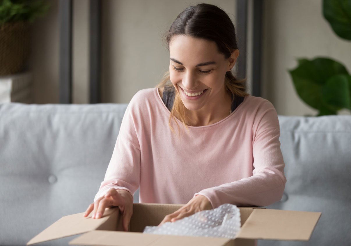 Mercado Pago: Uma mulher com blusa rosa rindo, enquanto abre uma caixa de papelão, que recebeu frete grátis