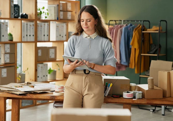 Mercado Pago: mulher em uma sala de estoque, vestindo calça bege, camisa branca com suéter azul por cima, mexendo no tablet para elaborar estratégias com ofertas de inverno para a empresa