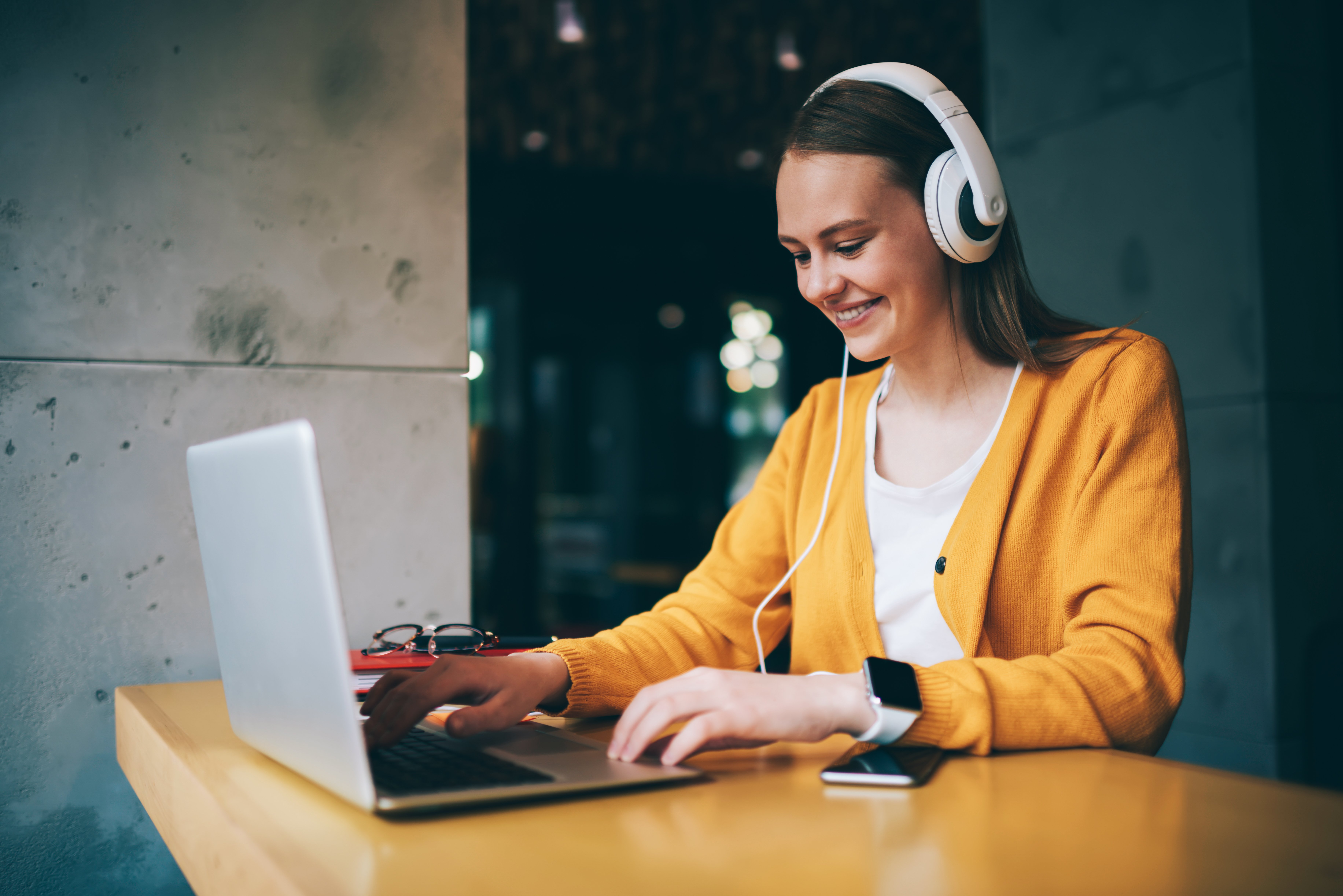 Mercado Pago e Bilheteria digital: Mulher com paletó amarelo e camiseta branca, com fone branco e sentada em mesa de escritório utilizando notebook.