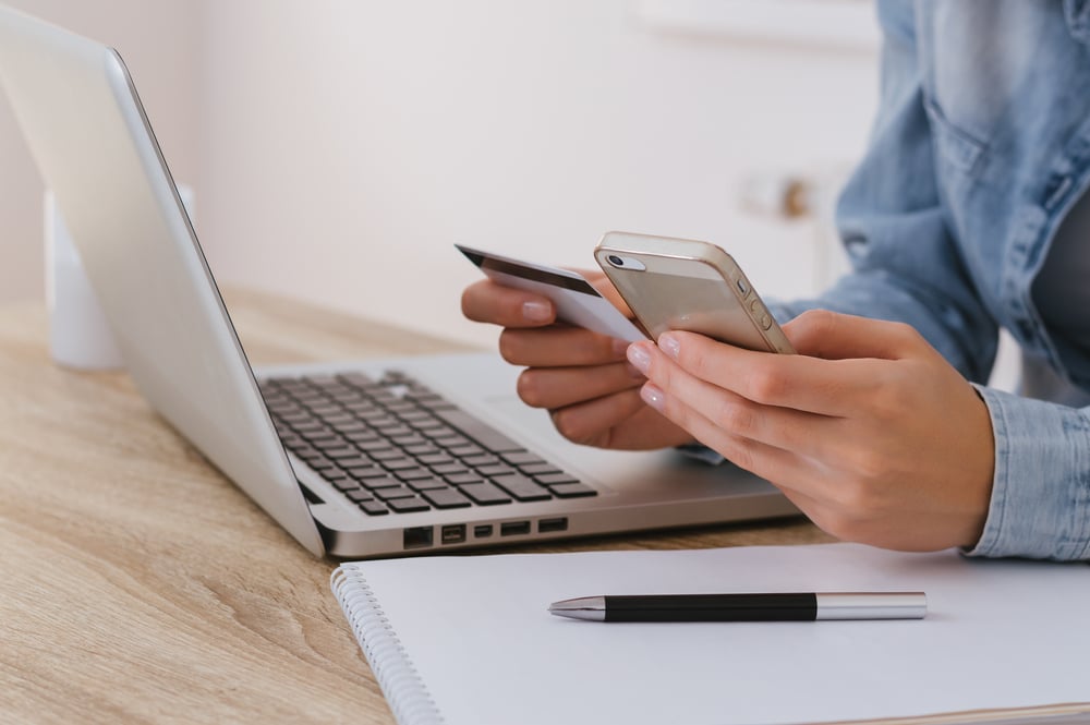 Mercado Pago: Mão segurando celular e cartão para realizar processo de pagamento em frente ao notebook, apoiado em mesa de escritório com caderno e caneta em cima também.